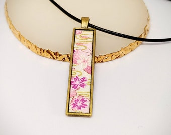 May include: A gold-toned pendant necklace with a rectangular charm featuring a pink and white floral pattern. The necklace is on a black cord.