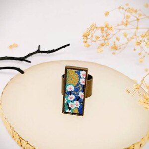 Op de afbeelding: Een messing ring met een rechthoekig blauw, goud en wit bloemenmotief. De ring ligt op een wit oppervlak met een gouden rand.