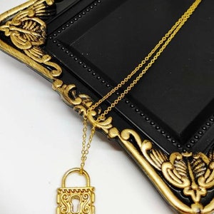 May include: A gold padlock necklace with an ornate design. The necklace is on a black tray with gold accents.