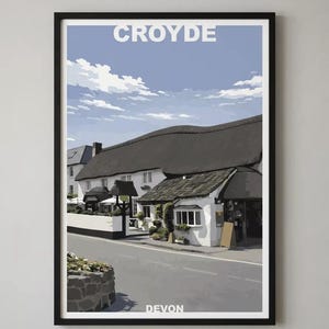 May include: Framed print featuring a stylized illustration of a building in Croyde, Devon. The artwork depicts a white building with a thatched roof under a blue sky with clouds. The word "CROYDE" is at the top, and "DEVON" is at the bottom.