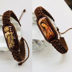 Peut inclure: Deux bracelets tressés marrons avec un charme à image religieuse. Un charme représente une femme en robe bleue, l'autre un homme en robe dorée.