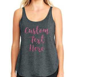 May include: A dark gray tank top with a scoop neck and a curved hemline. The tank top features the text "Custom Text Here" in a pink glitter font. The model is wearing black pants.