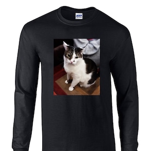 May include: A black long-sleeve t-shirt featuring a centered rectangular graphic print of a white and dark brown tabby cat sitting. The cat has a white face with dark markings around the eyes and ears, and a dark patch on its back. The shirt has a crew neckline.