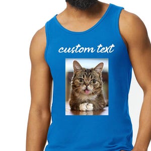 May include: A bright blue sleeveless tank top featuring a rectangular graphic print of a brown tabby cat with its tongue sticking out. Above the cat image, the words custom text are printed in a white, cursive, lowercase font style.