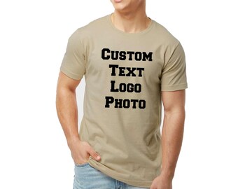 May include: A beige short-sleeved t-shirt featuring the black text CUSTOM TEXT LOGO PHOTO printed in a bold, block-style font on the center of the chest. The garment has a crew neckline and a standard fit, paired with light blue denim jeans.