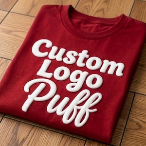 May include: A red t-shirt with the words "Custom Logo Puff" in white, raised lettering. The shirt is folded on a wooden surface. The design is centered on the front of the shirt.