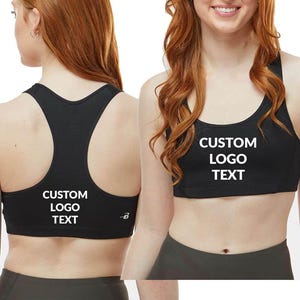 May include: Black sports bra with a racerback design. The bra features the text "CUSTOM LOGO TEXT" in white block letters on the front and back. The model is wearing dark gray leggings. The bra has a scoop neckline.