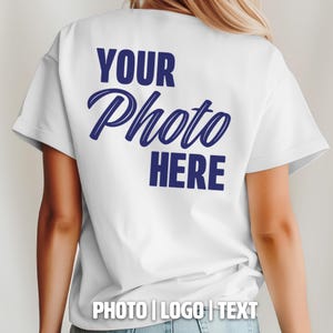 May include: White t-shirt with the text "YOUR Photo HERE" in navy blue script on the back. The bottom of the shirt has the text "PHOTO | LOGO | TEXT". The shirt is a basic crew neck.