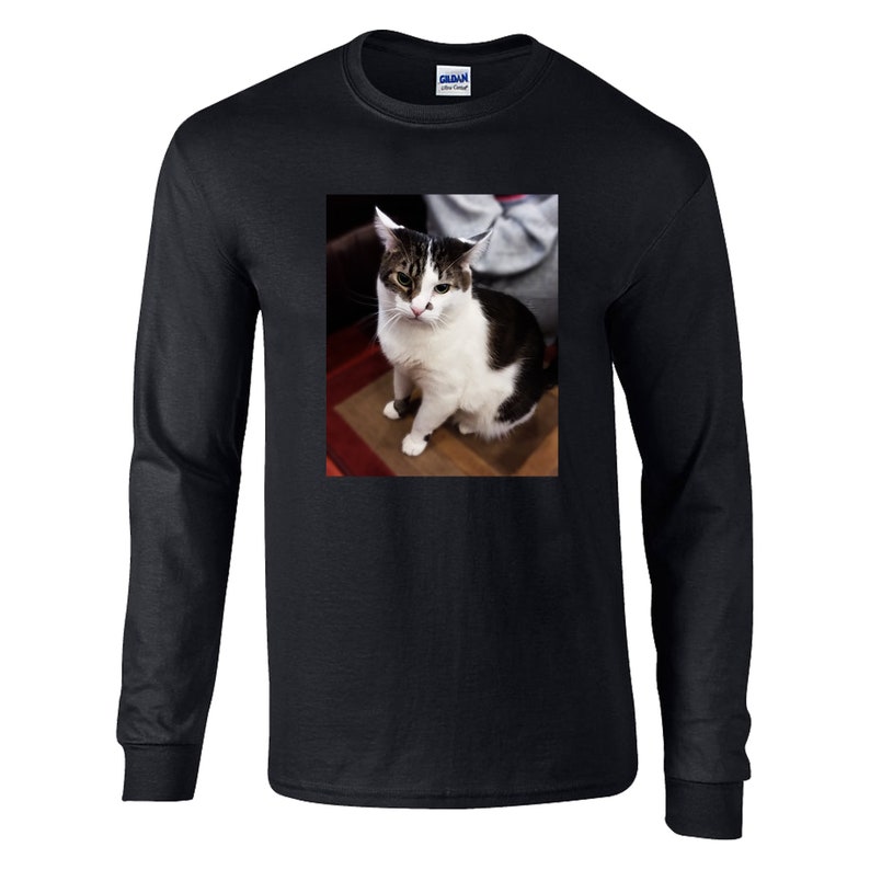 May include: A black long-sleeve t-shirt featuring a centered rectangular graphic print of a white and dark brown tabby cat sitting. The cat has a white face with dark markings around the eyes and ears, and a dark patch on its back. The shirt has a crew neckline.