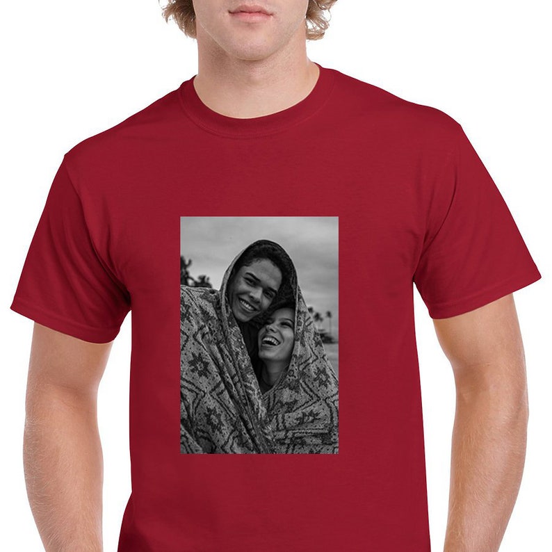 May include: A maroon t-shirt featuring a black and white photograph of two people smiling and wrapped in a patterned blanket. The photograph is centered on the shirt.