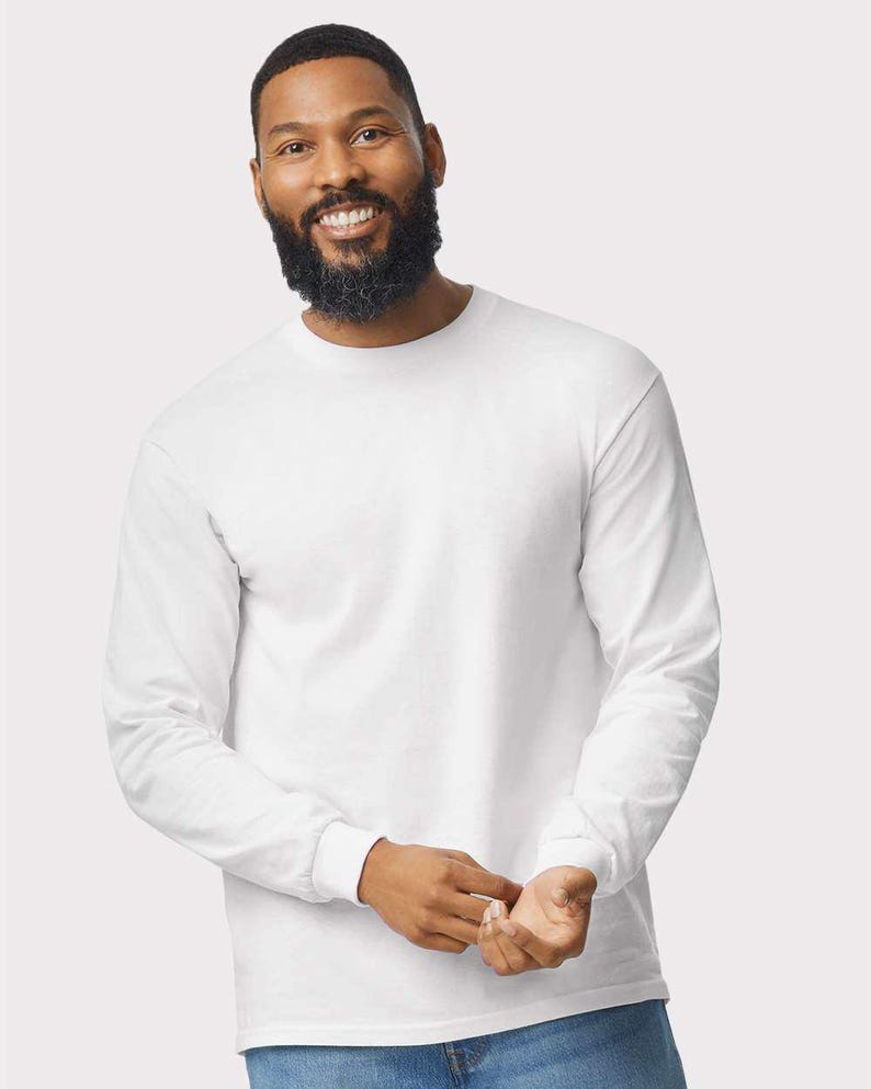 May include: A plain white long-sleeve t-shirt featuring a classic crew neckline and fitted cuffs at the wrists. The garment is constructed from a solid white fabric with a smooth texture, providing a clean and simple appearance for everyday wear.