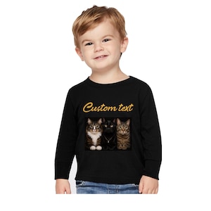 May include: A black long-sleeve shirt featuring the words Custom text in gold lettering above a rectangular graphic of three cats sitting side by side. The cats include a brown tabby, a solid black cat, and a brown tabby with stripes.