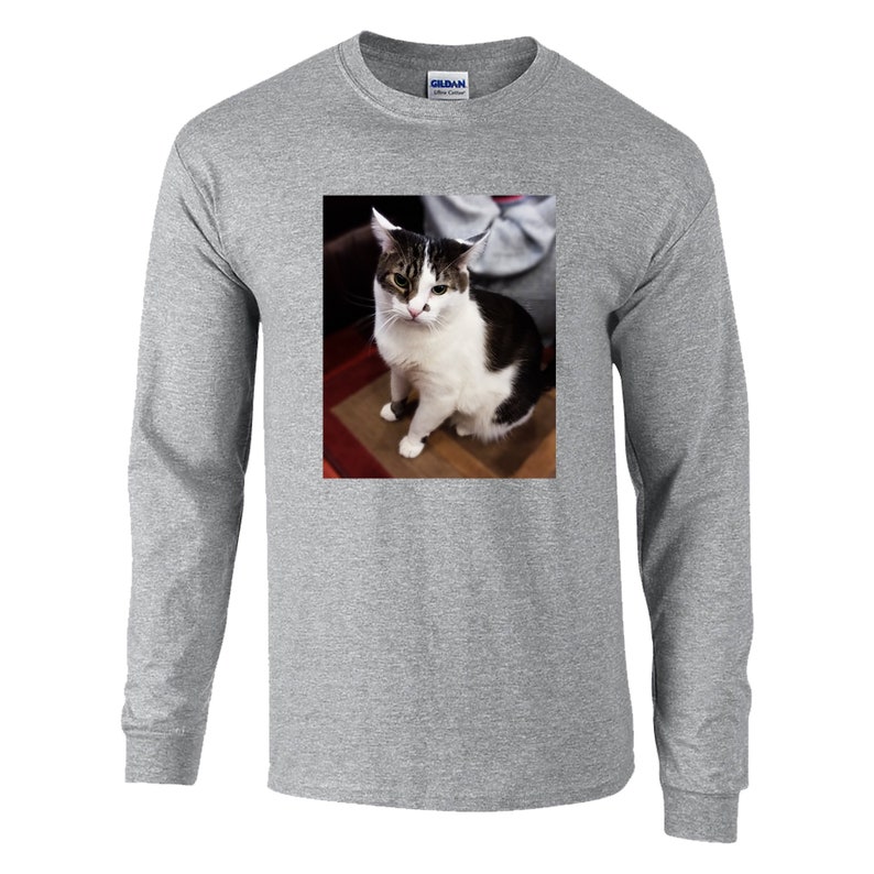 May include: A heather gray long-sleeve t-shirt featuring a centered rectangular graphic print of a white and dark brown tabby cat sitting down. The cat has a white face with dark brown markings around the ears and eyes, and a small dark spot on its nose.