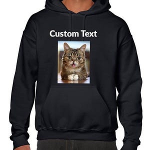 May include: A black hooded sweatshirt featuring a rectangular graphic print on the chest. The print displays a brown tabby cat with its tongue sticking out and the text Custom Text printed in white above the cat image on the black fabric.