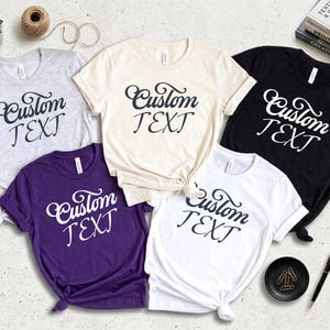 May include: A collection of five t-shirts in various colors: gray, cream, purple, white, and black. Each shirt features the words "Custom TEXT" in a stylish script. The shirts are neatly arranged, showcasing the design.