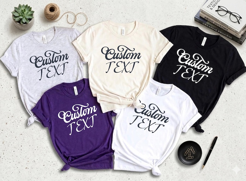 May include: A collection of five t-shirts in various colors: gray, cream, purple, white, and black. Each shirt features the words "Custom TEXT" in a stylish script. The shirts are neatly arranged, showcasing the design.