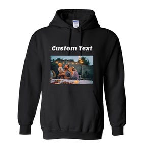 May include: A black hooded sweatshirt featuring a rectangular graphic print on the chest. The print displays a photograph of a family dining outdoors at dusk with string lights. Above the photograph, the words Custom Text are printed in white.