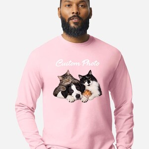 May include: A light pink long-sleeve shirt featuring a graphic print of a tabby cat, a black and white dog, and a small hamster huddled together. Above the animals, the text Custom Photo is printed in a white, cursive, script-style font.