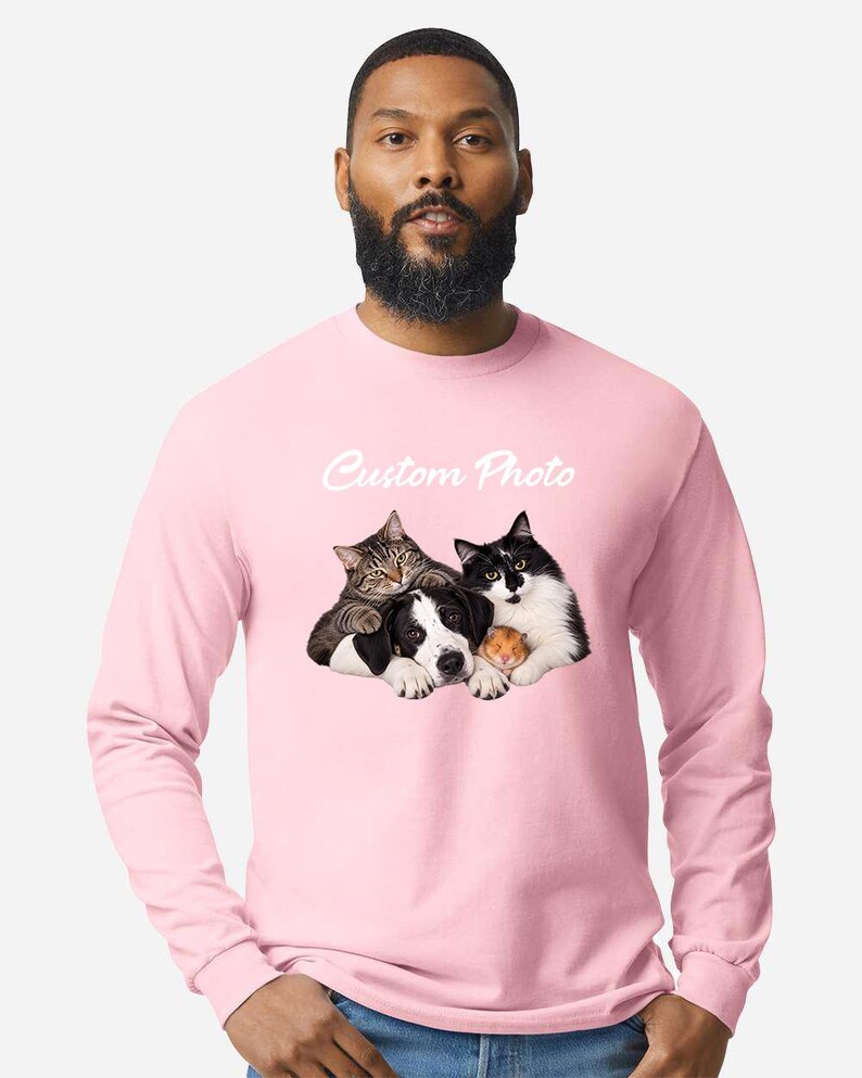 May include: A light pink long-sleeve shirt featuring a graphic print of a tabby cat, a black and white dog, and a small hamster huddled together. Above the animals, the text Custom Photo is printed in a white, cursive, script-style font.