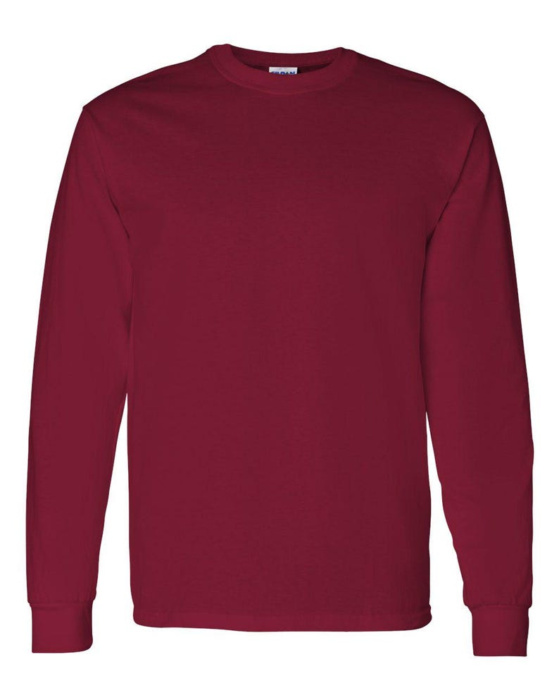 May include: A solid maroon long-sleeve crew neck t-shirt with a plain design. The garment features a standard ribbed collar and long sleeves with cuffs at the wrists. A small white brand label is visible on the inside of the neckline.