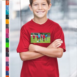 May include: A red youth-sized performance t-shirt with a graphic of a soccer team playing on a green field.