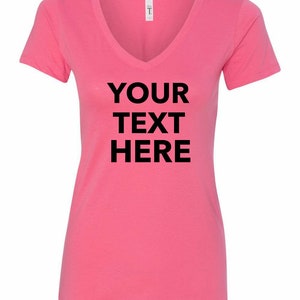 May include: A bright pink short-sleeved V-neck t-shirt featuring the black text "YOUR TEXT HERE" printed in a bold, sans-serif font across the center of the chest. The garment includes a white brand label at the inner neckline.