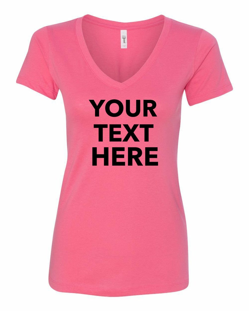 May include: A bright pink short-sleeved V-neck t-shirt featuring the black text "YOUR TEXT HERE" printed in a bold, sans-serif font across the center of the chest. The garment includes a white brand label at the inner neckline.