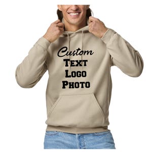 May include: A beige hooded sweatshirt featuring a front kangaroo pocket and long sleeves. The center of the chest displays the black text Custom TEXT LOGO PHOTO in a combination of script and block fonts. The garment includes a matching drawstring.