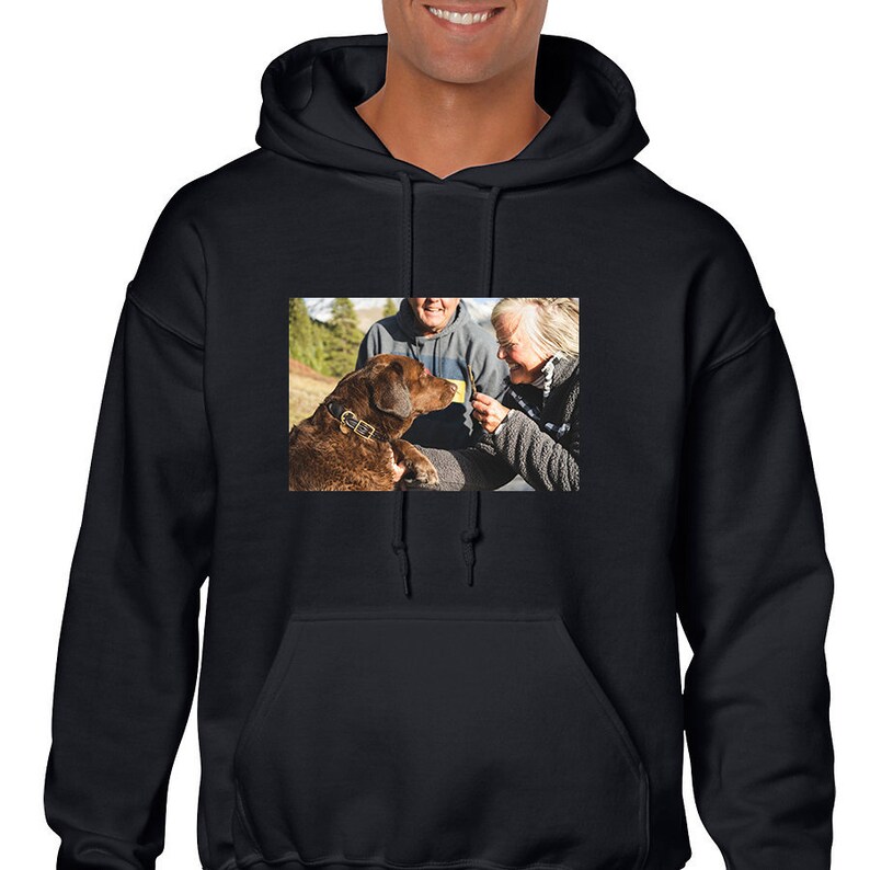 May include: A black hooded sweatshirt featuring a rectangular photographic print on the chest. The image displays a brown dog wearing a collar, positioned next to two individuals, with one person holding a small object toward the dog's face.
