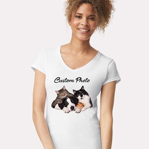 Op de afbeelding: Een wit T-shirt met V-hals voorzien van een grafische print van een cyperse kat, een zwart-witte kat, een zwart-witte hond en een kleine hamster die tegen elkaar aan liggen. De tekst Custom Photo staat in zwarte letters boven de illustratie.