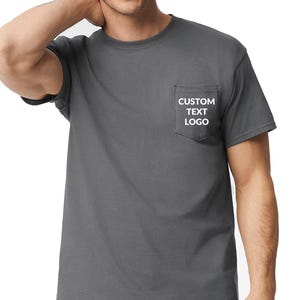 May include: A charcoal gray t-shirt with a pocket on the left chest. The pocket features the white text "CUSTOM TEXT LOGO". The shirt has short sleeves and a crew neck.
