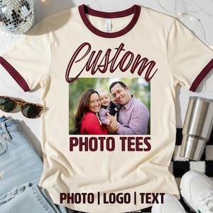 May include: Cream-colored ringer tee with burgundy trim featuring a family photo. The shirt has the word "Custom" in a script font above the photo, with "PHOTO TEES" and "PHOTO | LOGO | TEXT" below.