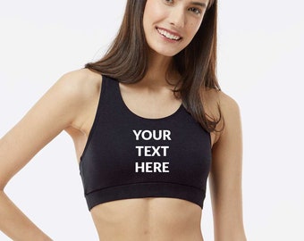 May include: Black sports bra with the text "YOUR TEXT HERE" in white. The bra has a scoop neckline and a racerback design. The model is wearing black bottoms.