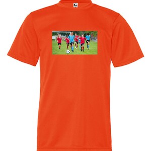 May include: Orange t-shirt with a graphic of a group of children playing soccer on a green field