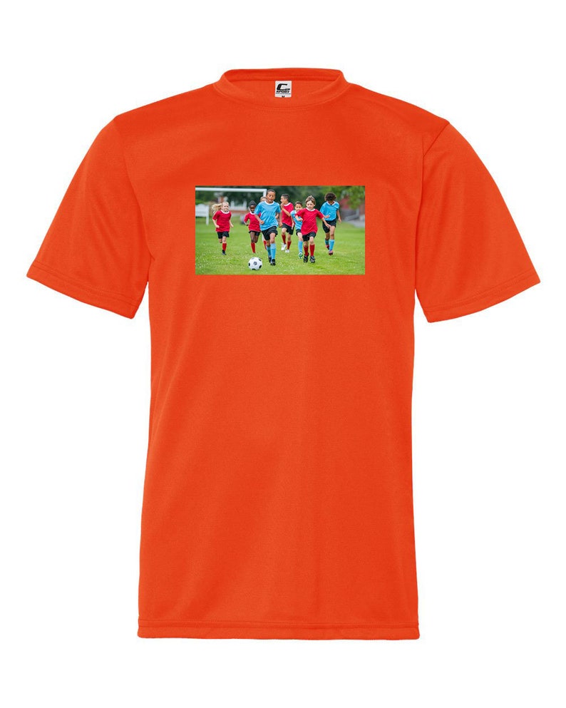 May include: Orange t-shirt with a graphic of a group of children playing soccer on a green field