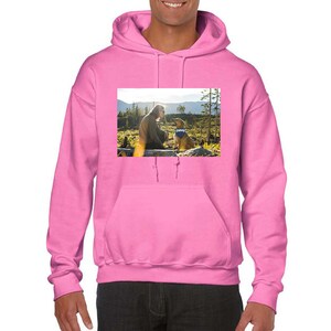 May include: A bright pink hooded sweatshirt featuring a rectangular graphic print on the chest. The print depicts a person kneeling in a forest landscape facing a dog wearing a blue bandana with the white text Chewy printed on it.