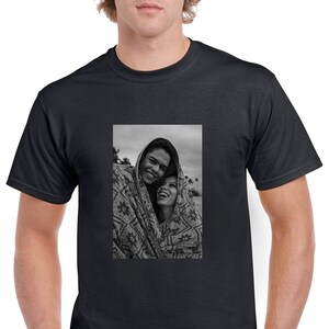 May include: Black t-shirt featuring a black and white image of two people smiling and wrapped in a patterned blanket. The image is centered on the shirt, showcasing a casual and comfortable style.