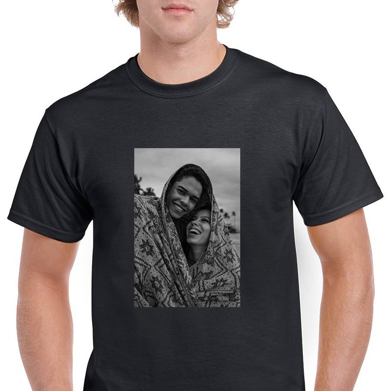 May include: Black t-shirt featuring a black and white image of two people smiling and wrapped in a patterned blanket. The image is centered on the shirt, showcasing a casual and comfortable style.