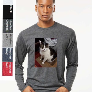May include: A long-sleeved t-shirt in a dark heather grey colour featuring a printed image of a tabby cat with white markings. The cat is sitting on a brown surface.