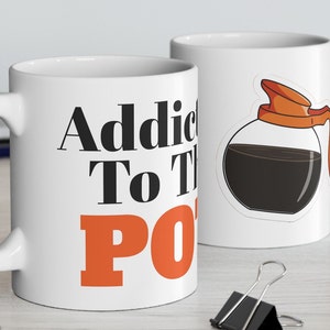 Può includere: Tazza da caffè in ceramica bianca con una grafica nera e arancione che dice "Addicted to the Pot" e un'immagine di cartone animato di una caffettiera.