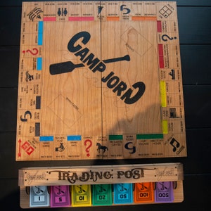 May include: A wooden board game called "Camp Jord" with a paddle and the game's name printed on the board. The game board has a variety of spaces with different prices and names, including "Trading Post", "Dining Hall", "Horse Stables", and "Program Center". The game also includes a wooden box with a drawer that holds the game's money, which is printed with the game's name and different denominations.