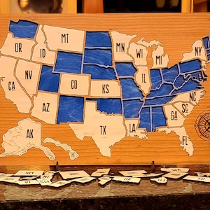 May include: A wooden map of the United States with each state outlined and labeled. The states are painted blue and the map is mounted on a wooden base. There are small wooden state pieces scattered on a dark surface.