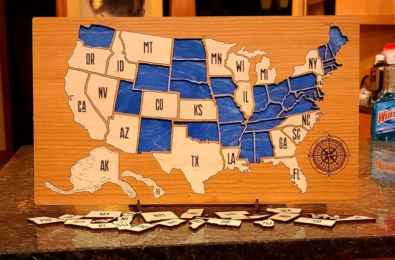 May include: A wooden map of the United States with each state outlined and labeled. The states are painted blue and the map is mounted on a wooden base. There are small wooden state pieces scattered on a dark surface.
