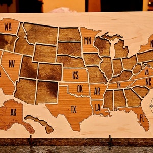May include: A wooden map of the United States with each state outlined and labeled. The map is stained in a dark brown color and has a compass rose in the bottom right corner.