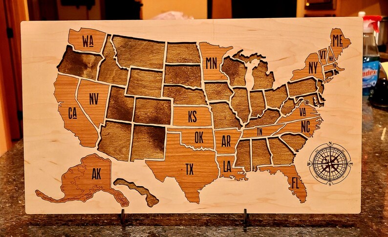 May include: A wooden map of the United States with each state outlined and labeled. The map is stained in a dark brown color and has a compass rose in the bottom right corner.
