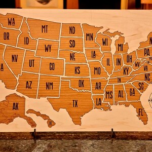 May include: A wooden map of the United States with each state outlined and labeled. The map is mounted on a stand and has a compass in the bottom right corner.