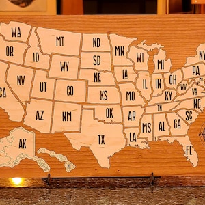 May include: A wooden map of the United States with each state outlined and labeled. The map is mounted on a wooden base with a compass in the bottom right corner.