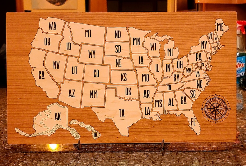 May include: A wooden map of the United States with each state outlined and labeled. The map is mounted on a wooden base with a compass in the bottom right corner.