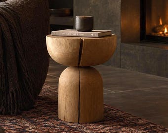 May include: A light brown wooden side table with a cylindrical base and a rounded top, featuring a vertical split design. A dark gray ceramic bowl and a book titled The Kinfolk Home are placed on the flat surface of the wooden table.