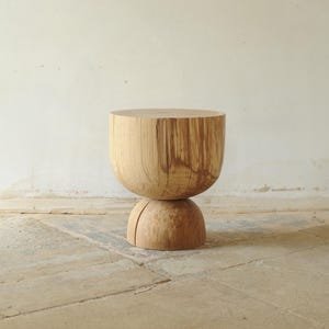 Puede incluir: Un taburete de madera hecho a mano con una parte superior redondeada y una base esférica. La veta natural de la madera es visible, mostrando la textura del material. El taburete mide aproximadamente 46 cm de alto y 38 cm de ancho.